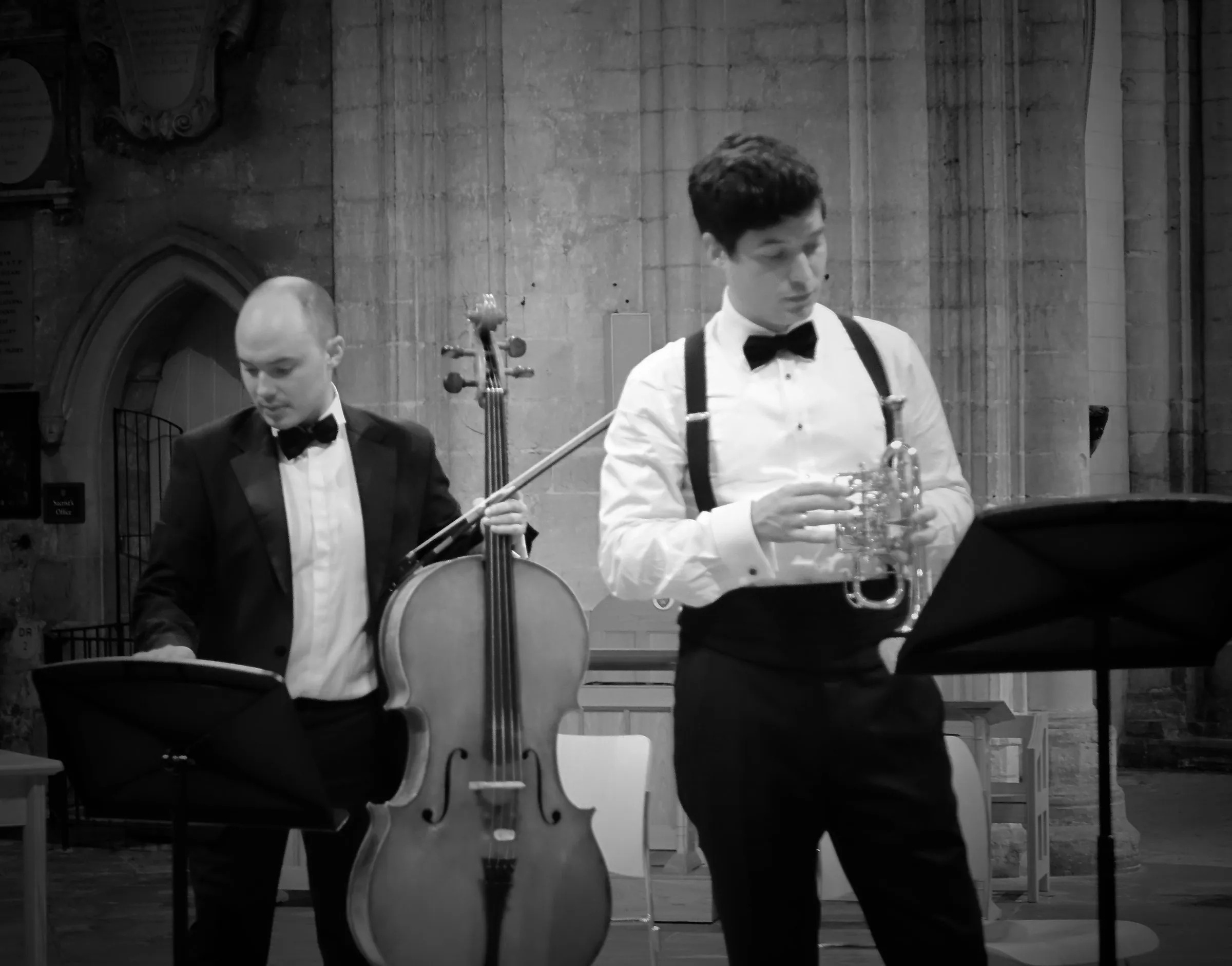 A cellist and a trumpeter in black tie at a music stand, in black and white