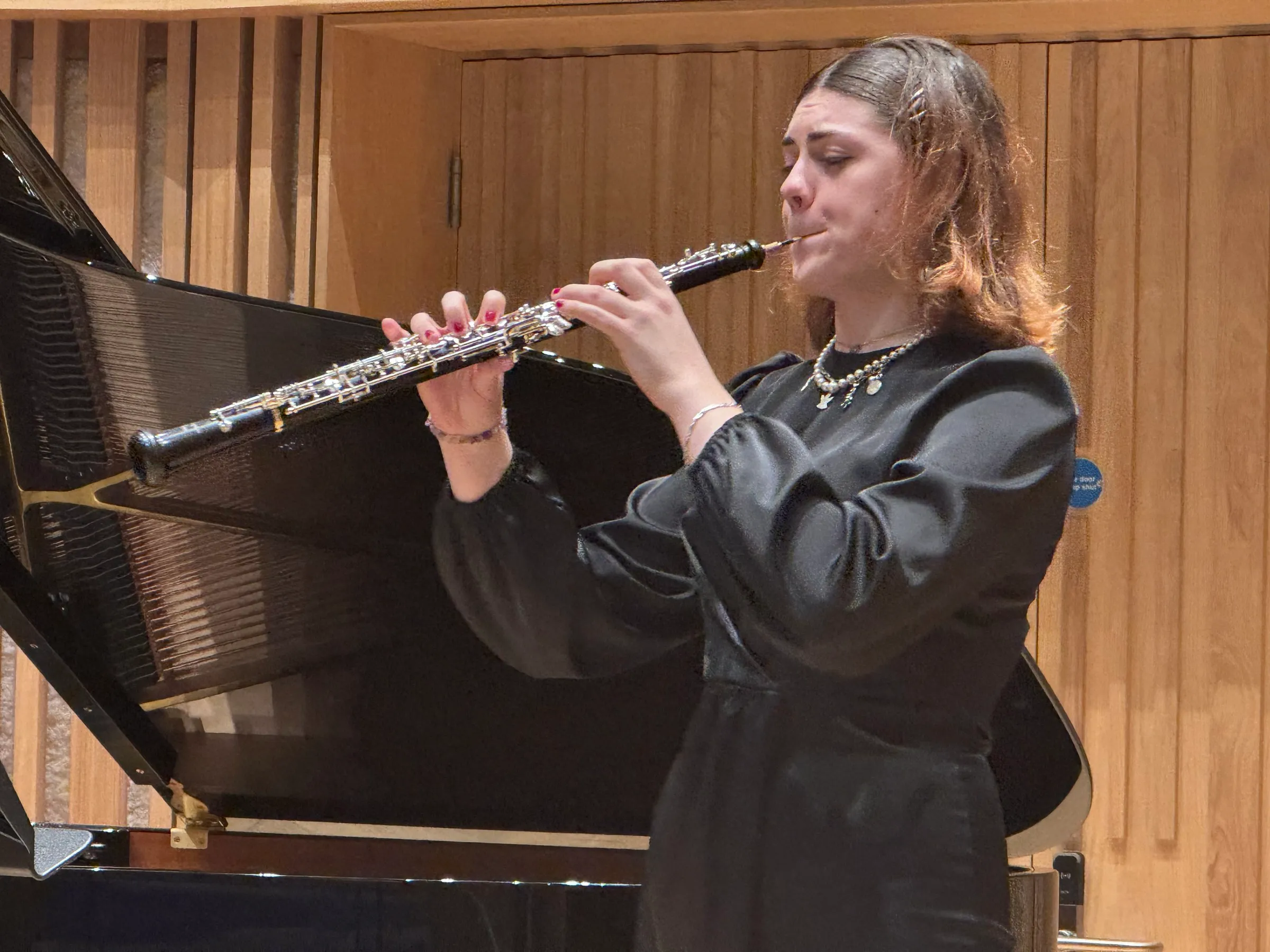 An oboist in a black dress plays beside the open grand piano