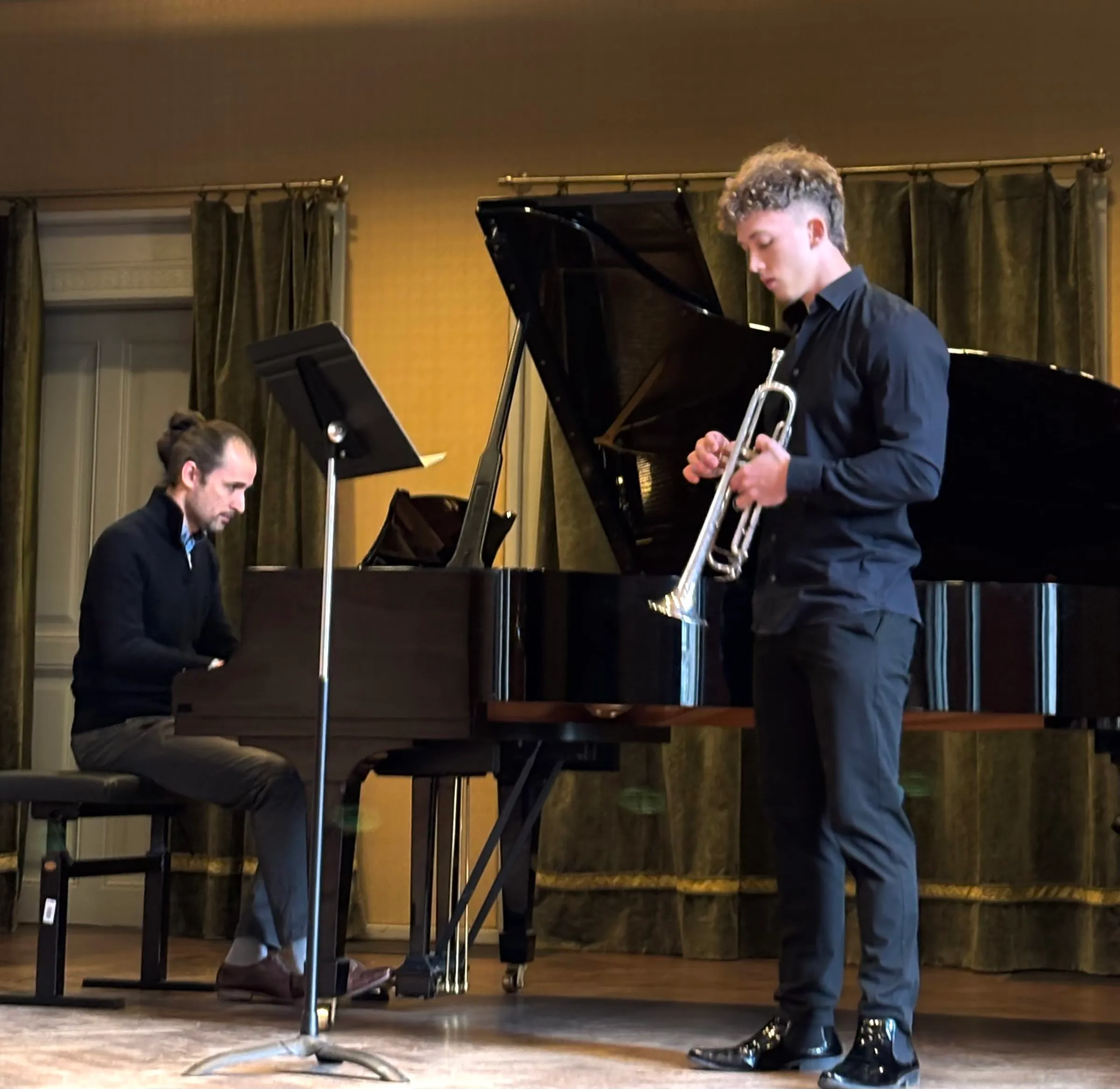 A trumpeter and pianist perform in front of green velvet curtains