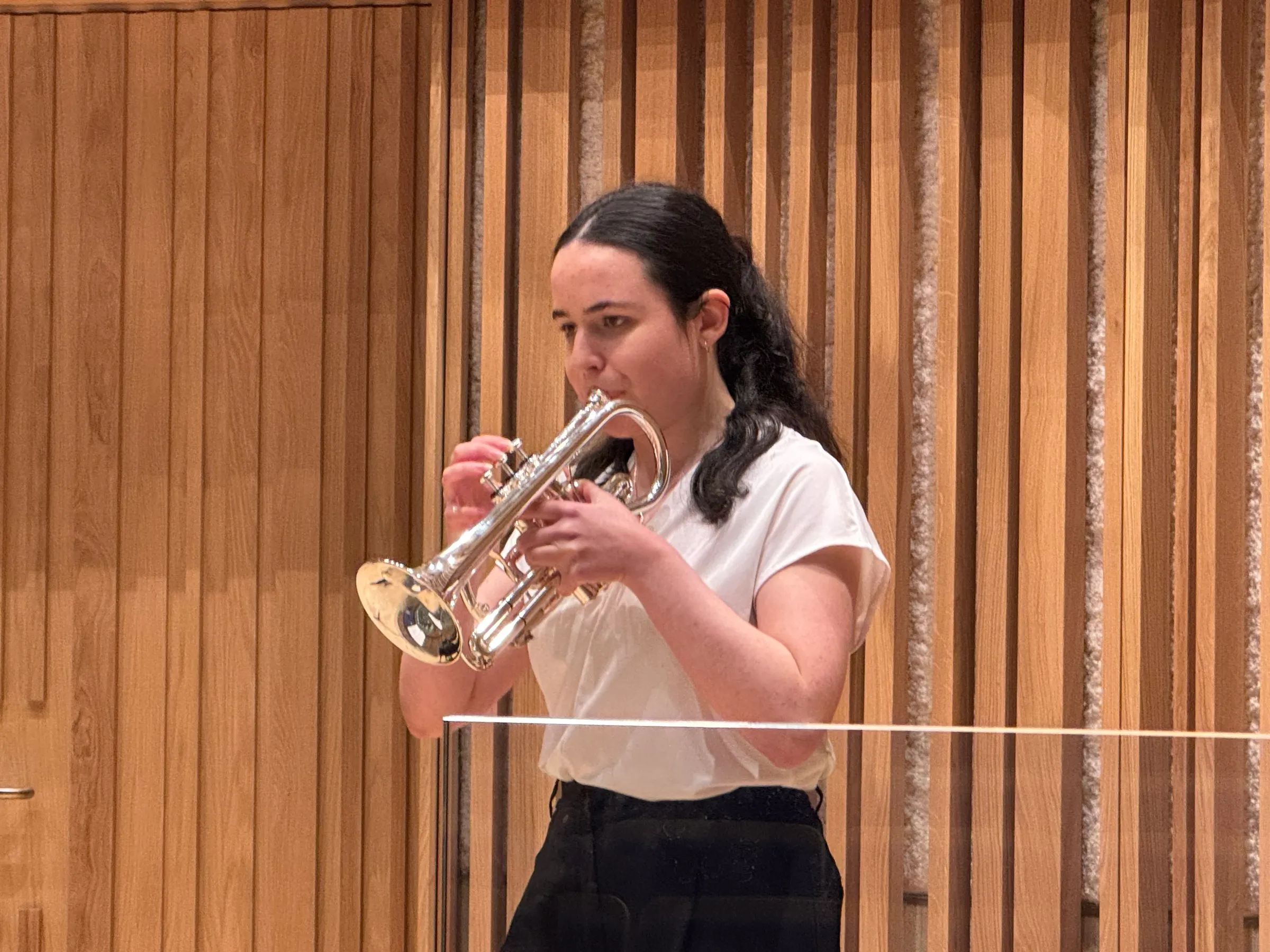 A young trumpeter in a white top plays in front of the hall's wooden slats
