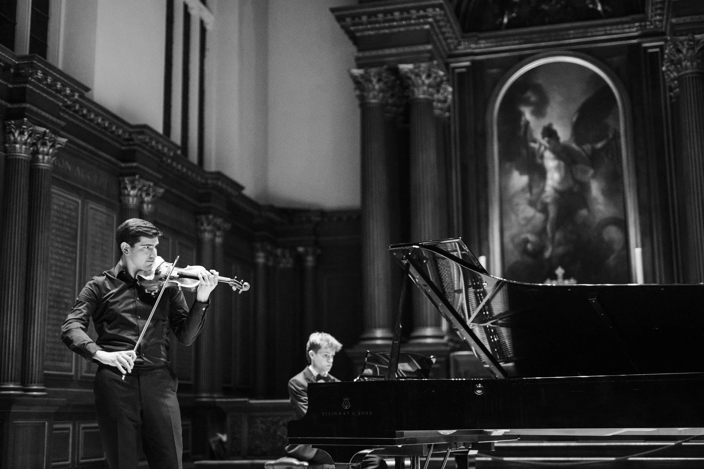 A violinist and pianist perform at the altar end of the chapel, in black and white