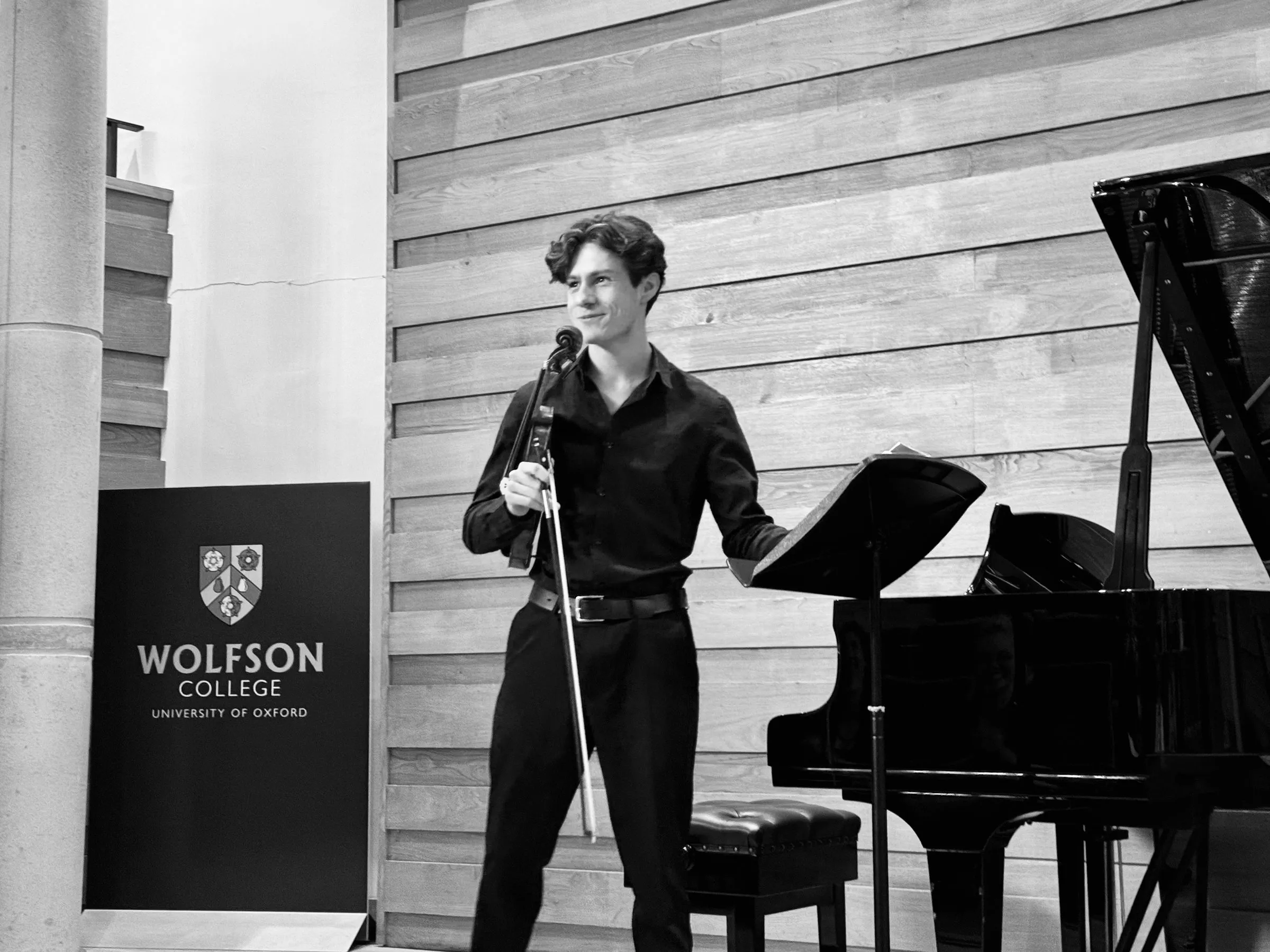 A violinist beside the Wolfson College banner and piano, in black and white